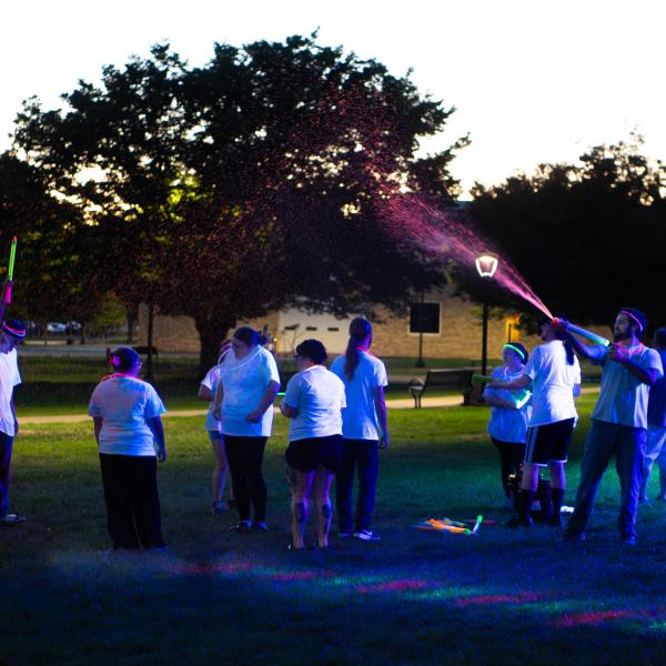 A student on the right sprays neon pink paint out a water blaster, over the heads of a group of fellow students.