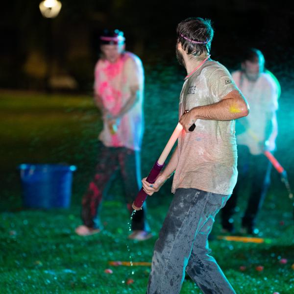 A student, whose clothing is soaked, holds a water blaster that has neon paint dripping from the end.