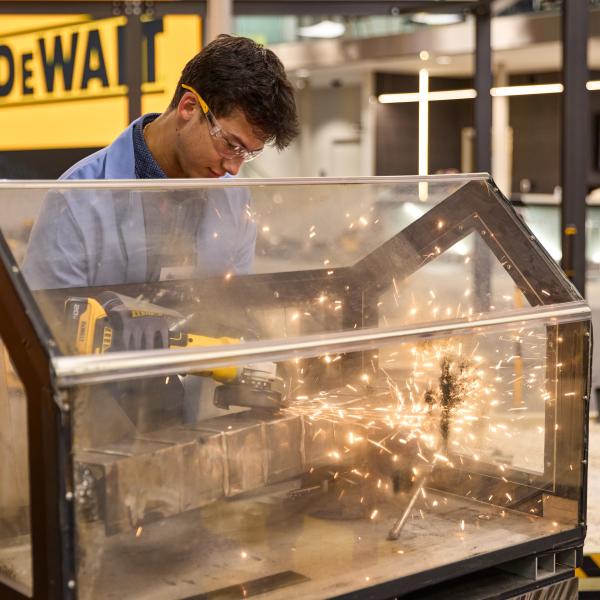 Sparks fly as a student wearing safety glasses and gloves runs a power tool against a block of metal. The tool and metal are inside a plexiglass enclosure.