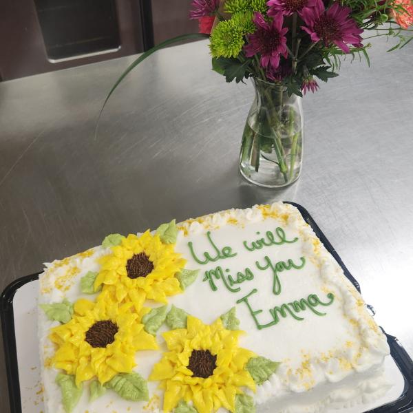 A vase full of flowers and a cake decorated with yellow flowers and the words "We will miss you, Emma!"