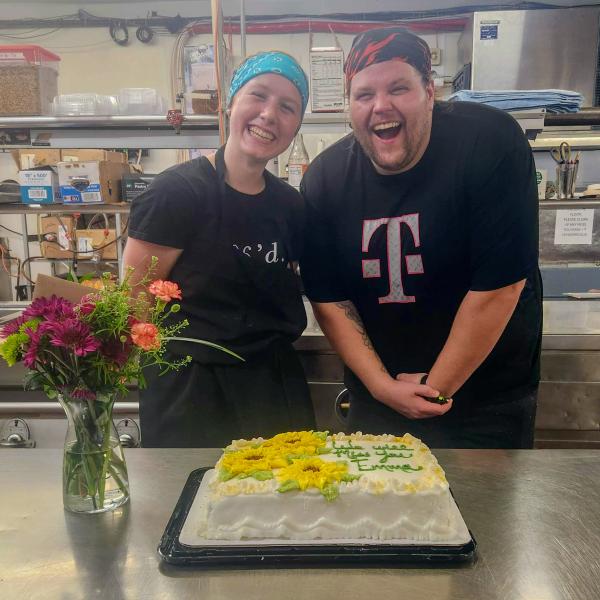 Two people smile widely while standing behind a table that has a cake and a vase of flowers.