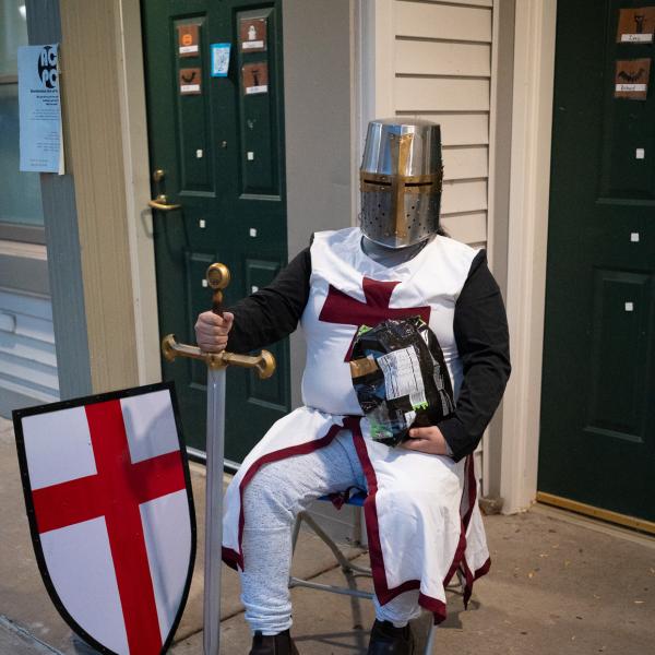 A student sits outside his apartment door with a bag of candy. He is earing a crusader costume.