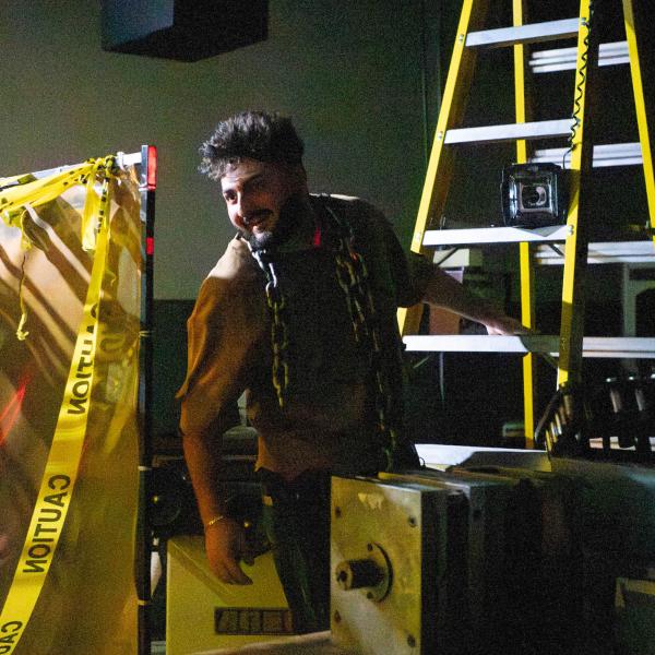 A student in a scary costume leans out between industrial equipment in a very dark room.