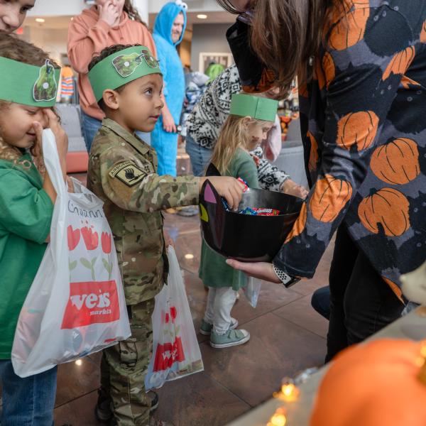 A woman holds a bowl of candy. A preschool-aged child in a camouflage costume looks at the woman as he reaches into the bowl.