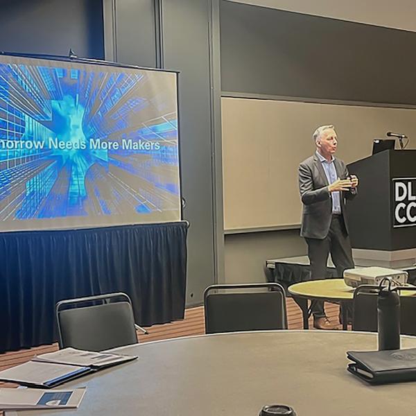 President Michael J. Reed speaks at the front of a conference room. Next to him is a screen that reads "Tomorrow Needs More Makers."