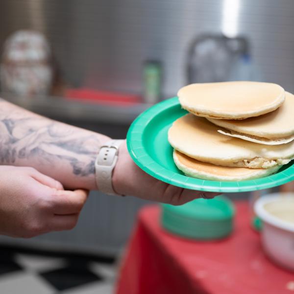 Person holding a green paper plate with a stack of pancakes in front of a table with pancake batter and other breakfast items.