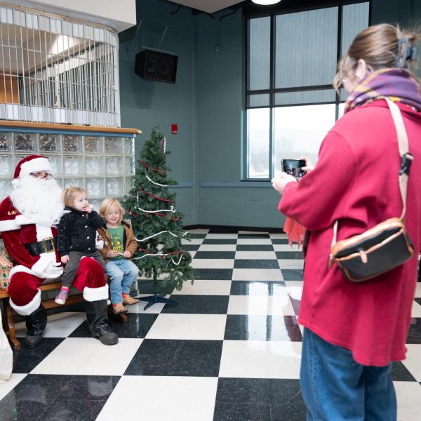 A woman holds up her phone to photograph two children sitting on Santa's lap.