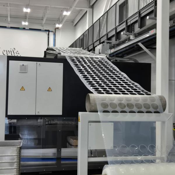 Industrial machine inside a manufacturing facility with a conveyor feeding a sheet of circular cutouts, and a roll of clear material in the foreground.