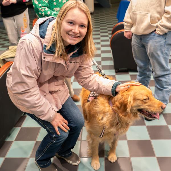 A person wearing a pink coat kneels beside a golden retriever, gently petting the dog as it stands on a tiled floor.