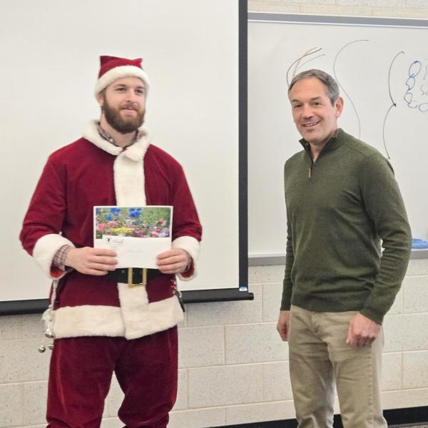 Two people stand in a classroom. One is wearing a santa suit and holding an award.