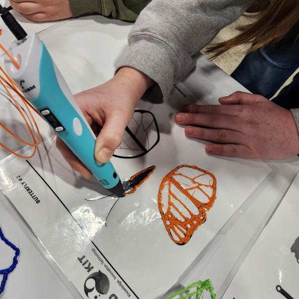 Close-up of hands guiding a teal 3D printing pen to draw an orange butterfly wing on a template sheet labeled for a 3D kit.