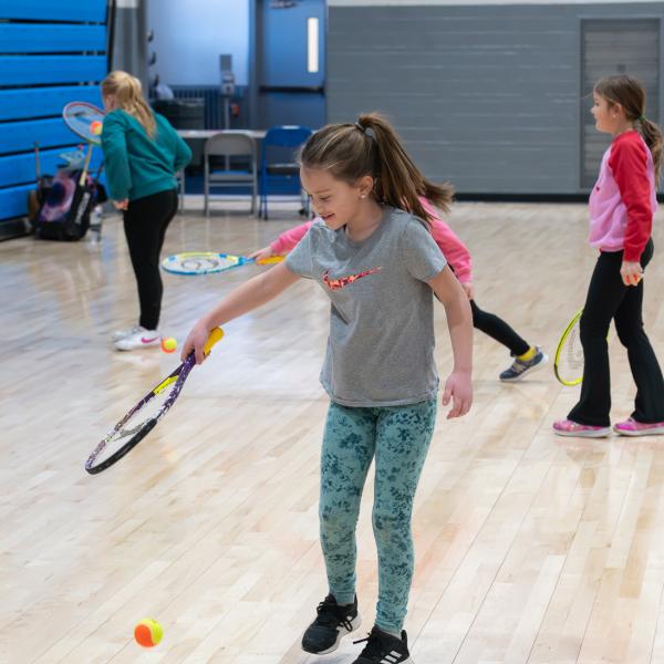 A girl uses a tennis racket to bounce a tennis ball on a gym floor.