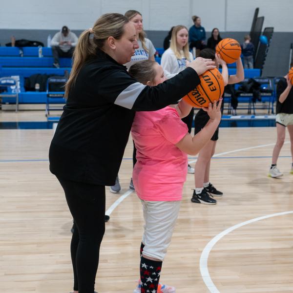 A woman helps a girl to aim a basketball toward a hoop.