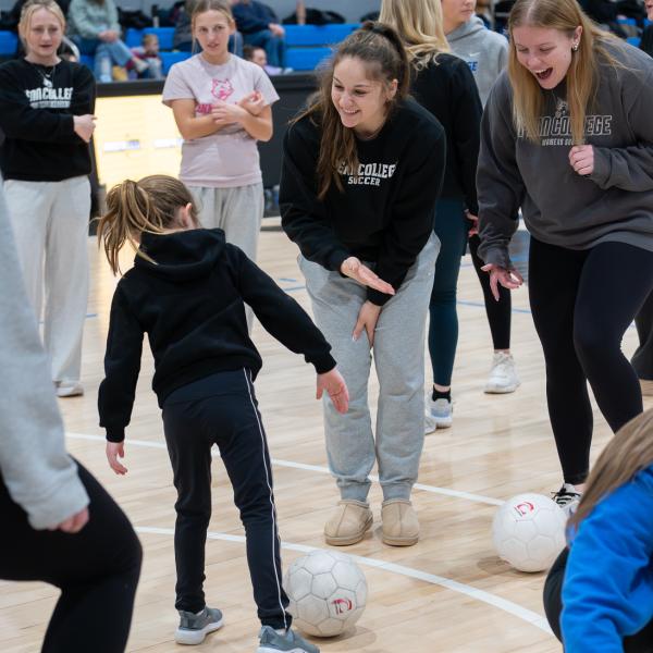 College athletes encourage a child as she dribbles a soccer ball.