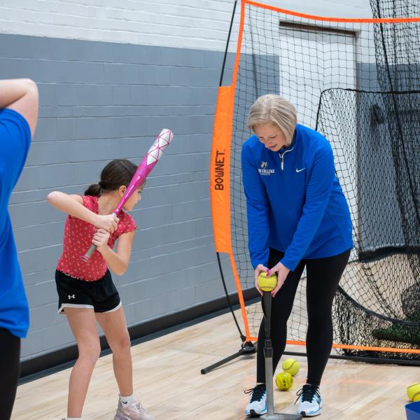 While a child waits with a bat, a woman places a softball on a tee.