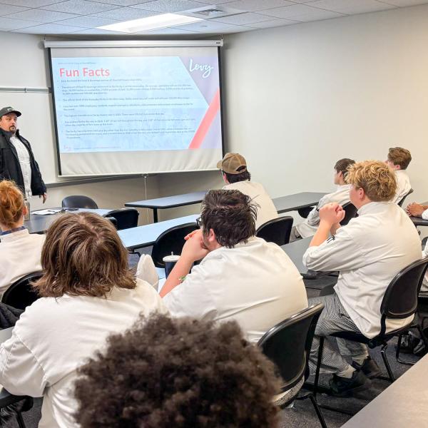 A man in a chef's jacket stands in front of a classroom of students sitting at desks wearing chefs uniforms.