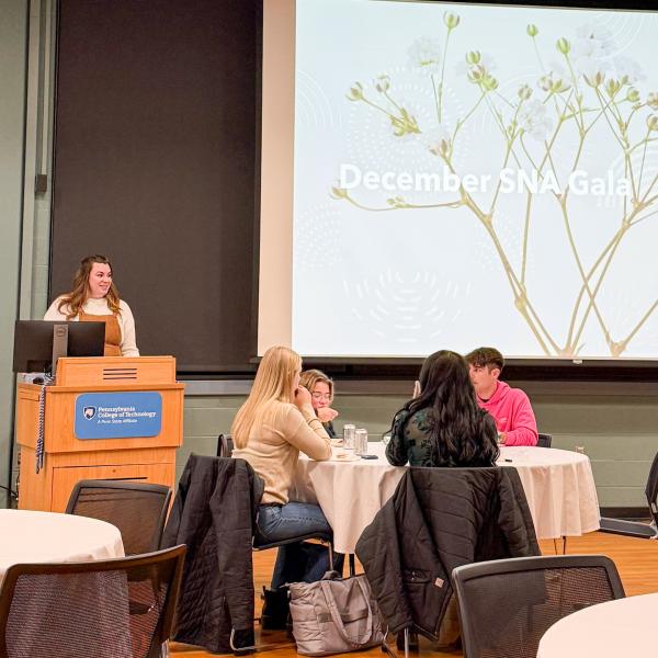 People seated at round tables look toward a student standing behind a podium in Penn College's Penn's Inn. A projection screen reads "December SNA Gala."