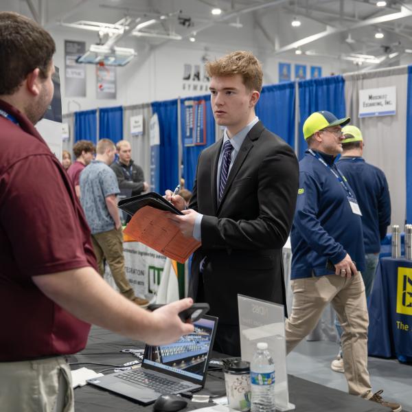 A student in a suit and tie holds a padfolio and takes notes while talking with an employer.