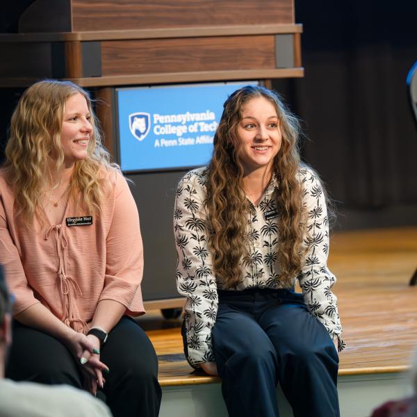 Two students sit on the edge of a stage, talking with people in the auditorium's seats.
