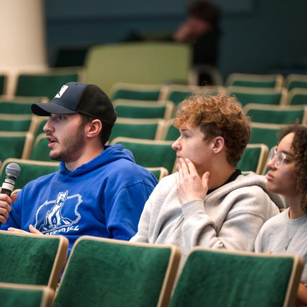 Three students sit in an auditorium. One is speaking into a microphone.