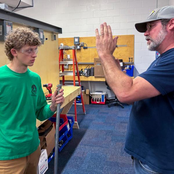 A student, holding a piece of conduit, looks at an instructor who is talking and motioning with his hands.