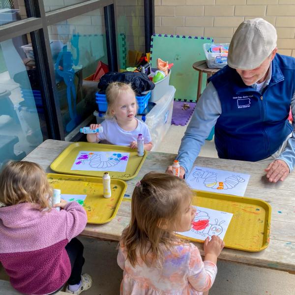 An adult sits with three children at a small picnic table. All are using dot paint markers to color pictures of a caterpillar.