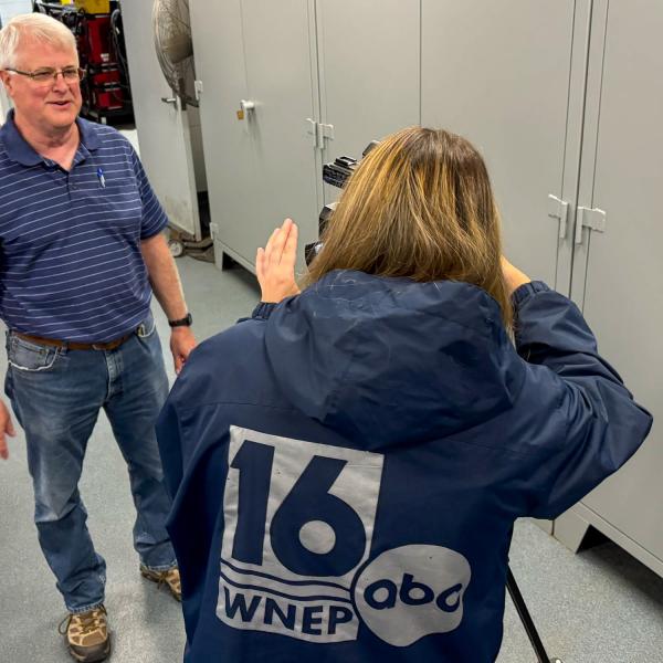 A man smiles as he stands in front of a television reporter who is operating a camera.