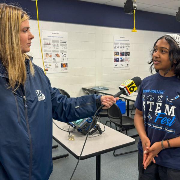 A television reporter holds a microphone up to a middle school student wearing a Penn College STEMFest T-shirt.