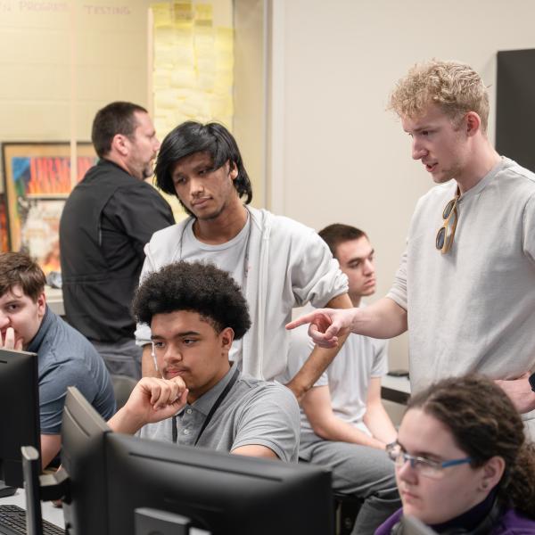Students sit behind computer screens while two others stand behind them, one pointing toward a screen.