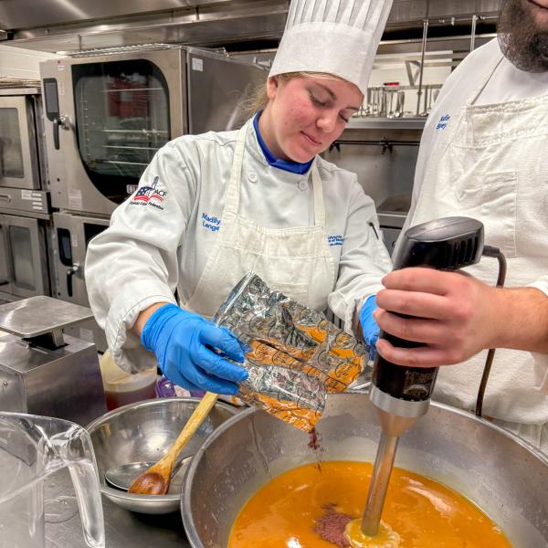 A student in a chef uniform adds ingredients to a large stainless steel bowl filled with liquid while another student in a chef uniform mixes it with an immersion blender.