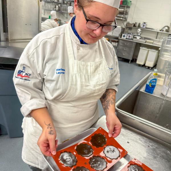 A student in a chef's uniform handles a silicone mold with six cavities that are filled with a brown liquid.