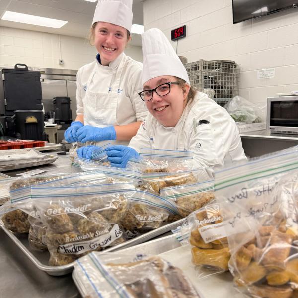 Student's in chef's uniforms add dog biscuits to zip-top plastic bags. Several bags of dog biscuits with hand-written labels sit in the foreground.