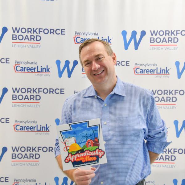 A man stands in front of a banner that says Pennsylvania CareerLink and Workforce Board Lehigh Valley and holds a small sign that says "Apprenticeship on the Move."