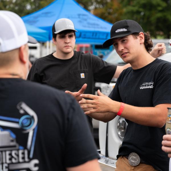Several people stand beside a vehicle at an outdoor automotive event. Gio Barbarossa, on right, gestures and speaks to the group. Blue canopy tents and parked vehicles are visible in the background.