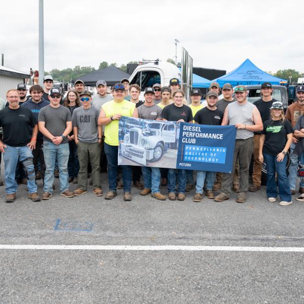 More than two dozen people pose in a parking area at Maple Grove Raceway, holding a banner for the Pennsylvania College of Technology Diesel Performance Club. Trucks, trailers, and event tents are visible in the background.