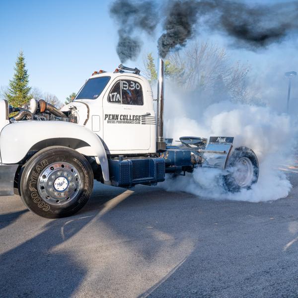 A white 1959 Mack semi-truck performs a tire burnout on a paved roadway at Penn College's Earth Science Center, producing thick white smoke around the rear wheels and black exhaust from vertical stacks.