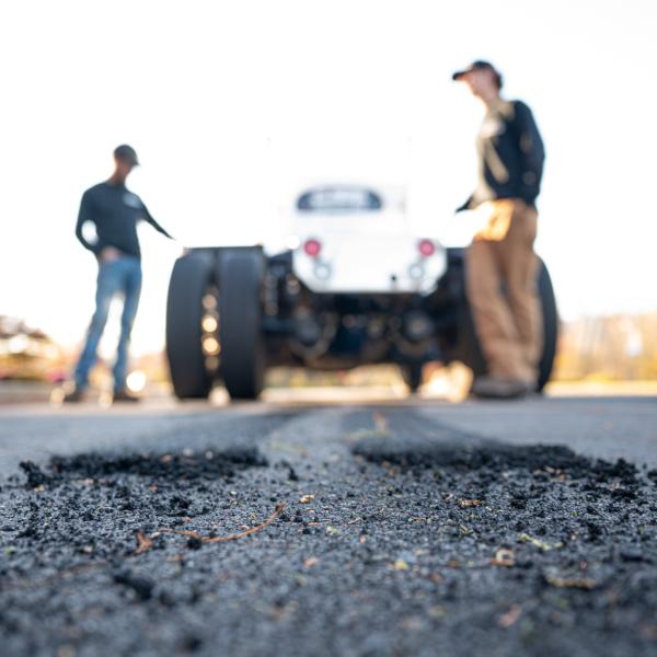 Close-up view of tire debris scattered on a roadway, with two people and a semi-truck blurred in the background. The image uses shallow depth of field to emphasize the worn pavement and rubber fragments in the foreground.