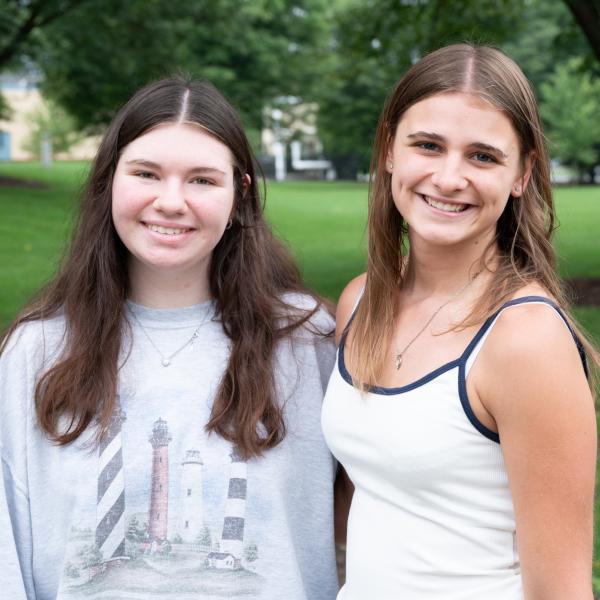 Two students smile at the camera. Trees and Penn College's Bush Campus Center are visible in the background.