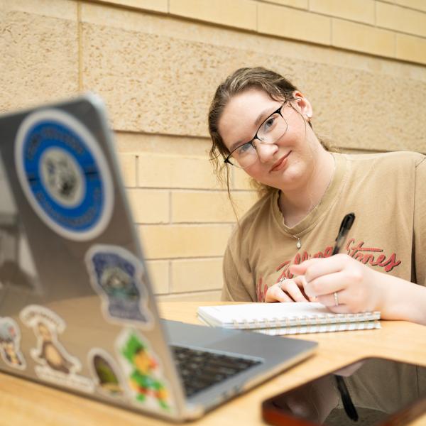 A student sits behind a laptop and writes in a small notebook while seated at a small table indoors.