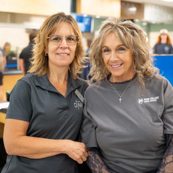 Two women stand close to each other and smile. Activity in the Keystone Dining Room is visible behind them.