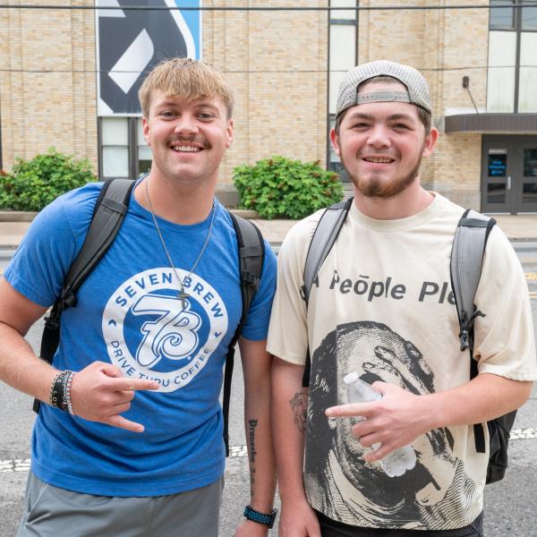 Two student make hand signals with their thumbs and pinkies extended. Penn College's Bardo Gymnasium is visible behind them.