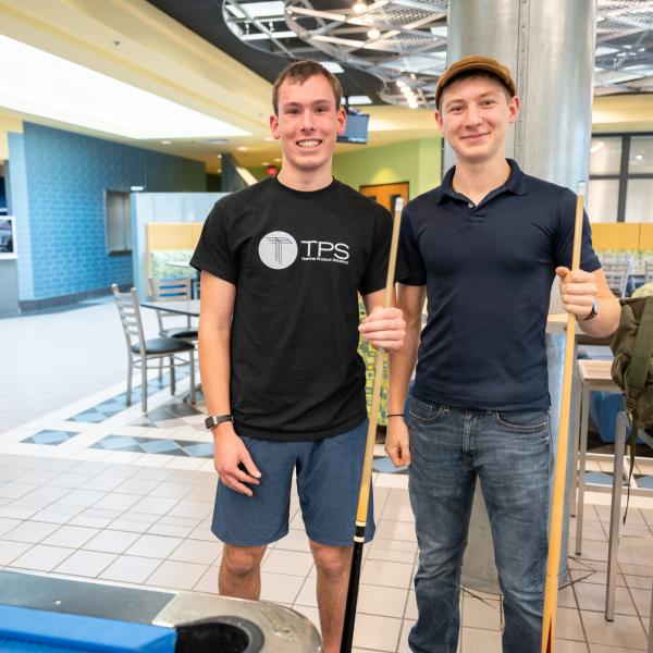 Two students hold pool sticks in the Bush Campus Center. The corner of a pool table is visible in the foreground.