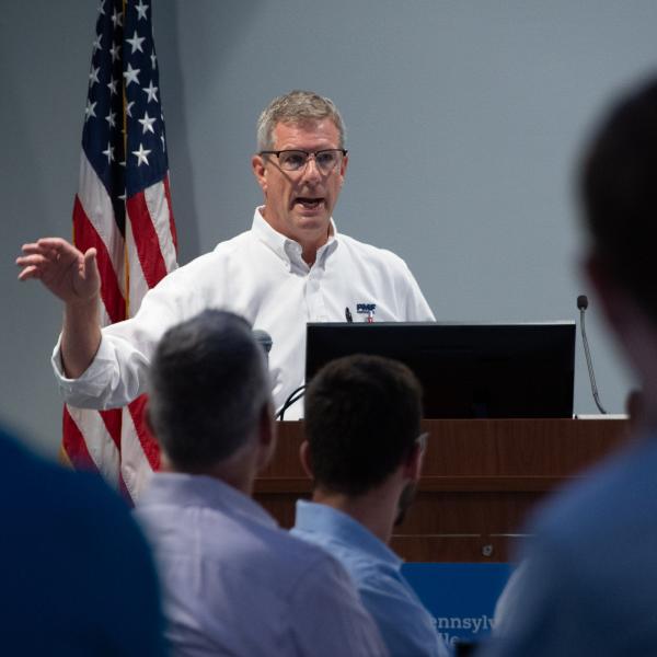 A man stands behind a microphone and speaks to a group of people seated in the foreground.