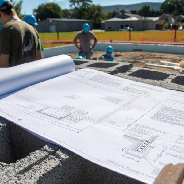 Blueprints sit open on a stack of concrete blocks while students in hard hats work in the background.