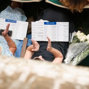 Two pairs of hands hold event programs. A bouquet of white roses sits in an empty auditorium seat.