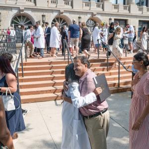 Families gather outside the Klump Academic Center. In the center, a woman in a long white lab coat and a man hug.