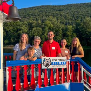 A family of six stands behind decorative red and blue railing- on the second floor of a riverboat - with the water, a green mountain and blue sky behind them. They hold a Penn College Alumni sign.
