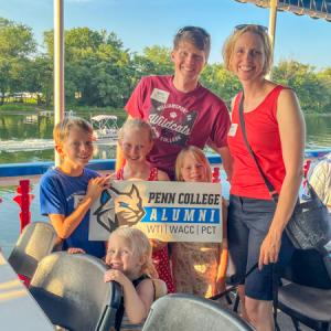 Two adults and four young children stand near table and chairs and hold a Penn College Alumni Relations sign. Behind them are water, a smaller boat, trees and blue sky.
