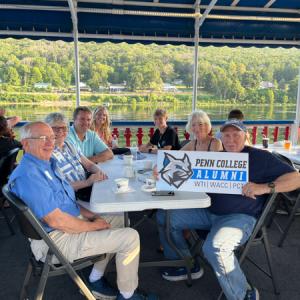 Seven people of varying ages sit at a table and hold a Penn College Alumni sign. Water and trees are in the background.