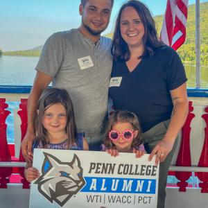 Two adults stand behind two young children holding a Penn College Alumni sign. Behind them are a red and blue railing, water, a hillside and blue sky.